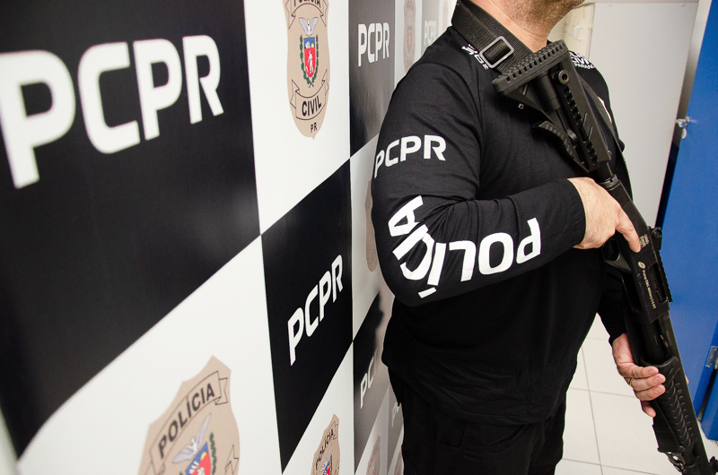 Policial civil empunhando arma ao lado de banner da PCPR