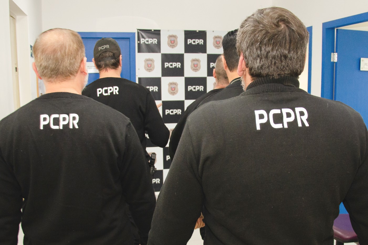 Vários policiais civis de costas. Ao fundo, banner da PCPR