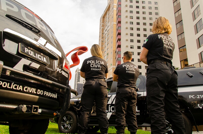 Três policiais civis de costas entre viaturas
