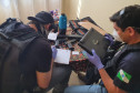 Dois policiais civis sentados, com objetos apreendidos sobre uma mesa.