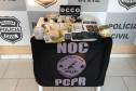 Materiais apreendidos, sobre uma mesa. Ao fundo, banner da polícia civil