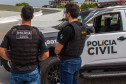 Dois policiais de costas, ao lado de viatura