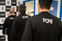 Três policiais civis de costas. Ao fundo, banner da PCPR