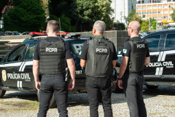 Tres policiais civis de costas próximos a viaturas
