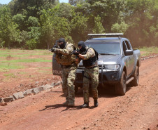 Dois policiais civis empunhando fuzis, andando lado a lado, em frente à viatura estacionada