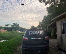 Policial civil ao lado de viatura do COPE e helicóptero sobrevoando o local