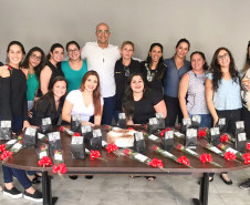 Quatorze mulheres posando para foto junto ao delegado, atrás de uma mesa com bolo e diversas rosas vermelhas