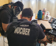 Dois policiais civis sentados, com objetos apreendidos sobre uma mesa.