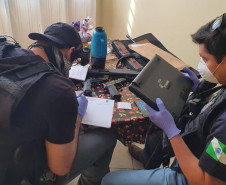 Dois policiais civis sentados, com objetos apreendidos sobre uma mesa.
