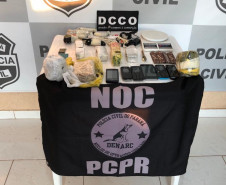 Materiais apreendidos, sobre uma mesa. Ao fundo, banner da polícia civil