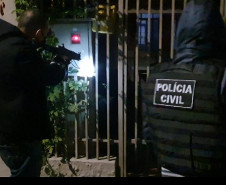Dois policiais civis em frente a um portão. Um deles aponta uma arma.