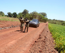 Policiais andando lado a lado apontando as armas, em frente à viatura em estrada rural