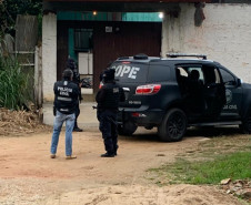 Policiais civis ao lado de viatura, em frente à uma residência