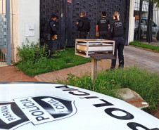 Policiais civis em frente a uma residência. Em primeiro plano, capô de viatura com brasão da polícia civil