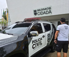 Policial civil ao lado de viatura, em frente à delegacia