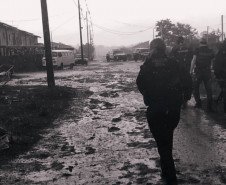 Policiais civis de costas, andando por via de terra após chuva