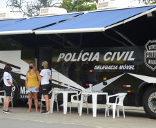 Duas policiais civis atendendo uma veranista ao lado do ônibus com a delegacia móvel