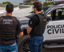 Dois policiais de costas, ao lado de viatura