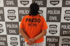 Mulher presa, voltada para uma parede com banner da Polícia Civil