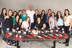 Quatorze mulheres posando para foto junto ao delegado, atrás de uma mesa com bolo e diversas rosas vermelhas