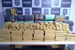 Drogas apreendidas empilhadas sobre uma mesa. Ao fundo, banner da Polícia Civil