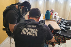 Dois policiais civis sentados, com objetos apreendidos sobre uma mesa.