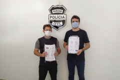 Policiais civis segurando embalagens com as máscaras. Ao fundo, brasão da Polícia Civil.