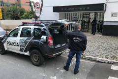 Policial em pé, segurando um par de algemas, ao lado de viatura com porta-malas aberto, em frente à delegacia