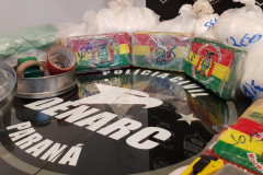 Material apreendido, sobre uma mesa. Ao centro, logomarca da DENARC