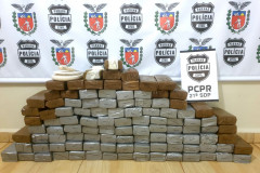 diversos tabletes da droga apreendida, empilhados no chão. Ao fundo, banner da polícia civil.