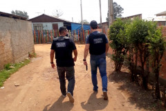 Dois policiais de costas, andando lado a lado, em rua de terra