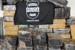 Tabletes de droga empilhados. Sobre eles, bandeira com logo da polícia civil
