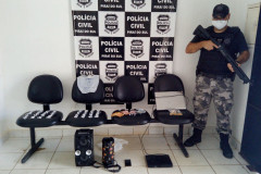 Policial civil empunhando arma ao lado de material apreendido. Ao fundo, banner da polícia civil.