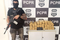 Policial empunhando arma ao lado de droga apreendida. ao fundo, banner da PCPR.