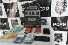 Material apreendido, sobre uma mesa. Ao fundo banner e logo da polícia civil
