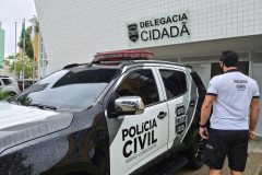 Policial civil ao lado de viatura, em frente à delegacia
