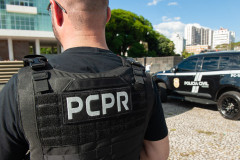 Policial civil de costas. Ao fundo, viatura.