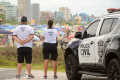 Dois policiais civis de costas, ao lado de viatura