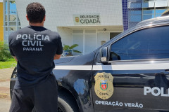 Policial civil de costas, ao lado de viatura em frente à Delegacia Cidadã