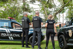 Três policiais civis de costas entre viaturas