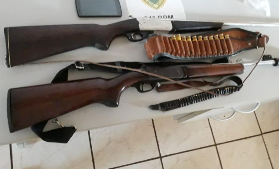 Duas armas, munição e um celular apreendidos, sobre uma mesa