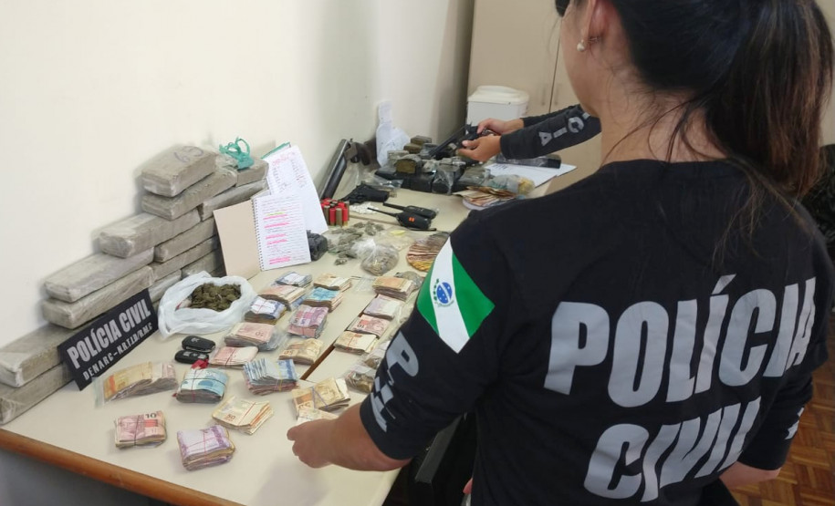 Policiais civis organizam, sobre uma mesa, drogas, armas, dinheiro e produtos apreendidos