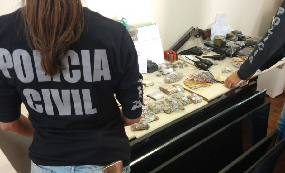 Policiais civis organizam, sobre uma mesa, drogas, armas, dinheiro e produtos apreendidos