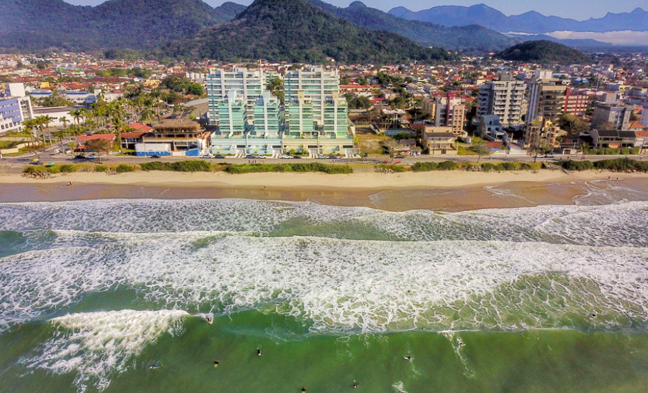 Litoral paranaense, com mar em primeiro plano e, ao fundo, prédios e montanhas.
