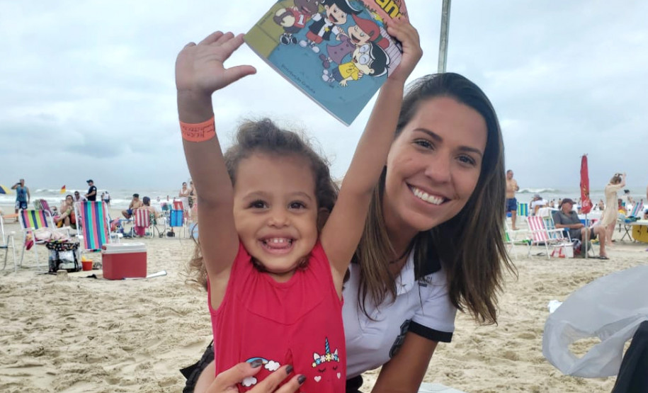 Policial civil abraça criança com pulseirinha, segurando revista da PCPR