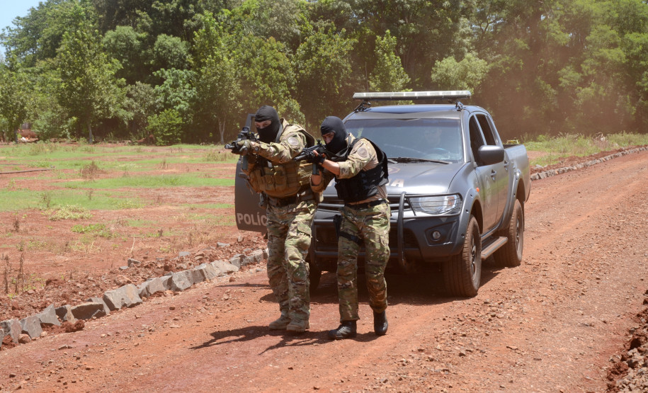 Dois policiais civis empunhando fuzis, andando lado a lado, em frente à viatura estacionada