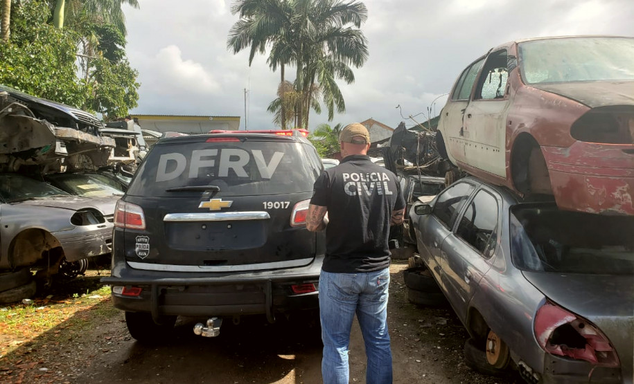 Policial civil ao lado de viatura, entre pilhas de carros desmanchados