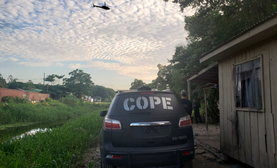 Policial civil ao lado de viatura do COPE e helicóptero sobrevoando o local