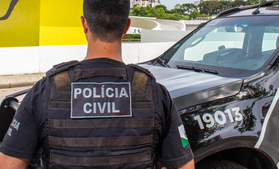 Policial civil, de costas, ao lado de viatura