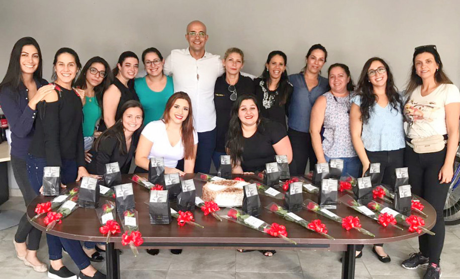 Quatorze mulheres posando para foto junto ao delegado, atrás de uma mesa com bolo e diversas rosas vermelhas
